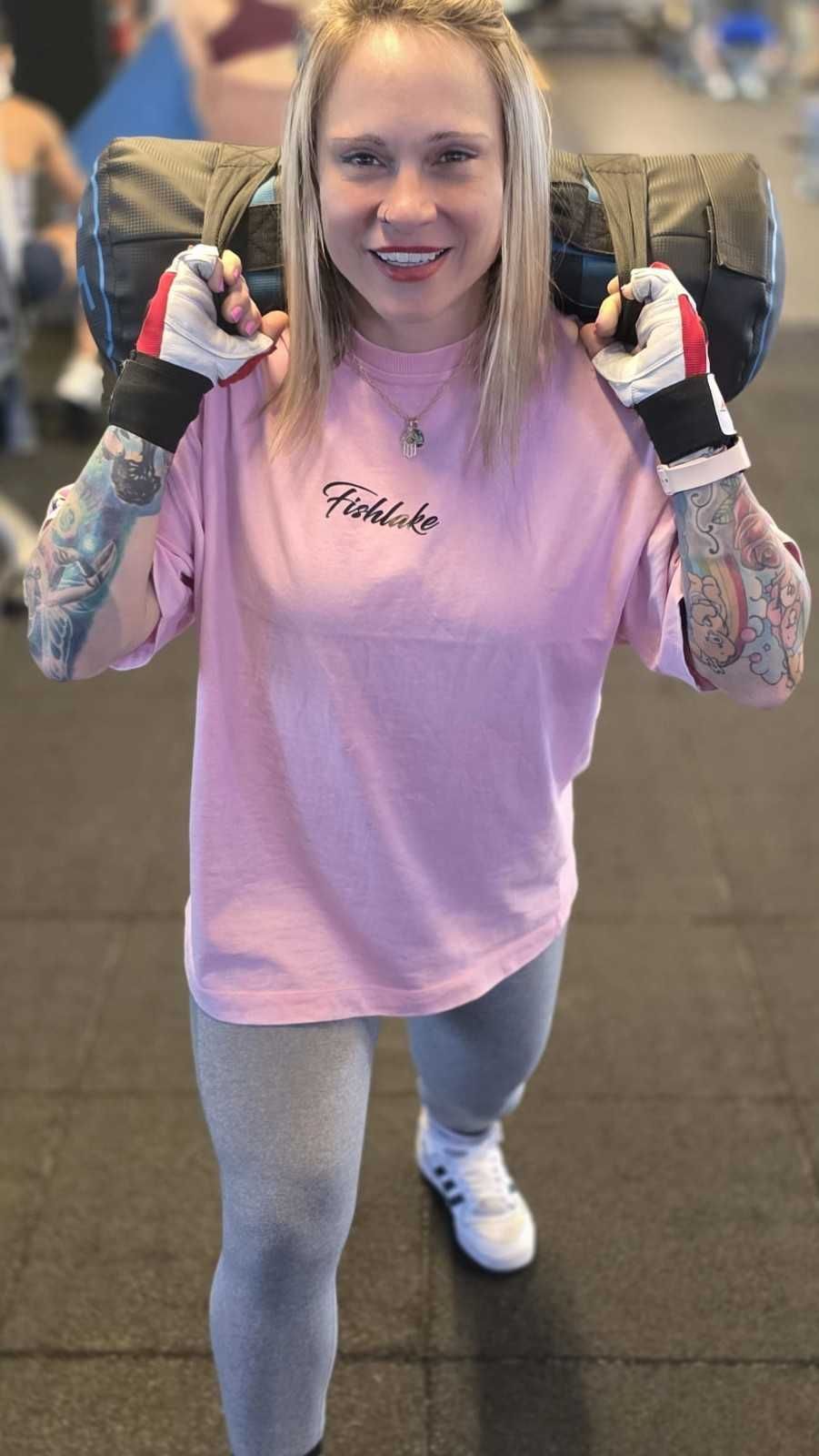 Persona sonriente con camiseta rosa y zapatos blancos, posando con los puños en alto en un gimnasio.