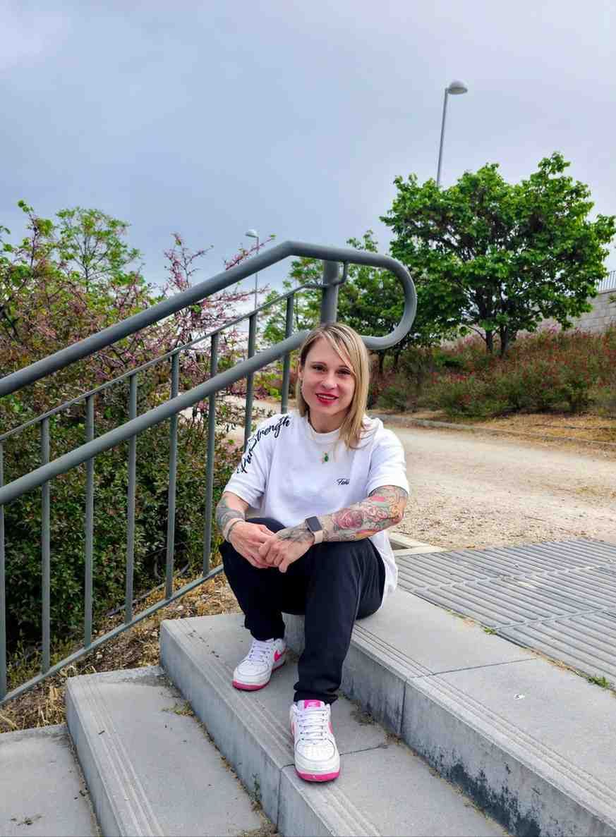 Persona sentada en unos escalones junto a una barandilla al aire libre, vestida con una camisa blanca, pantalones negros.