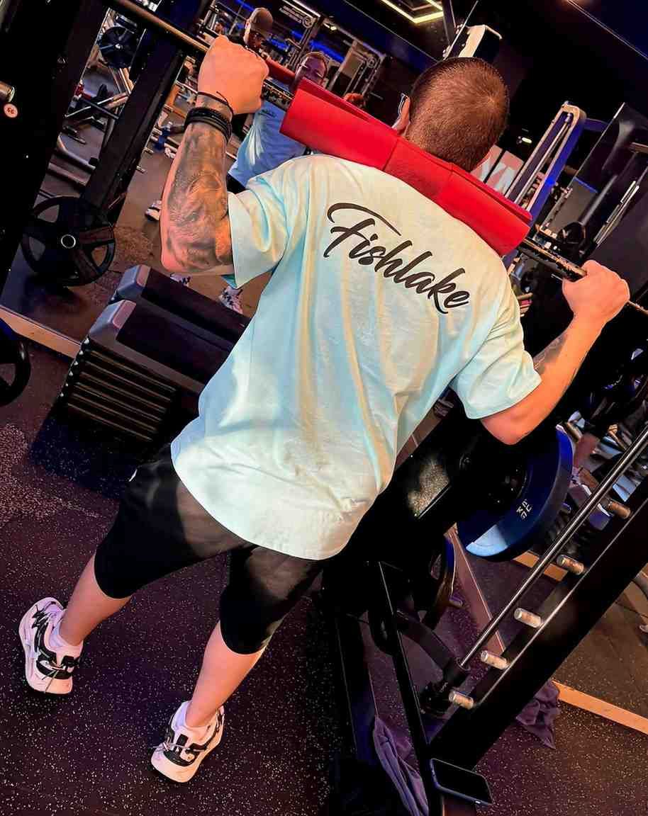 Persona levantando una barra en un gimnasio, vestida con una camiseta azul claro, pantalones cortos negros.