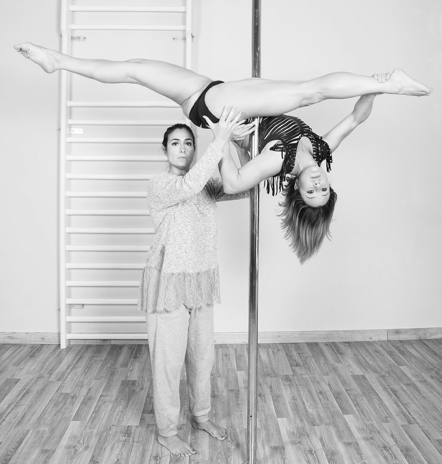 Mujer en pole en split, sostenida por otra en un estudio de baile.
