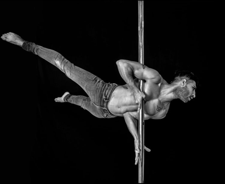 Un hombre con jeans hace una pose desafiante de pole dance sobre un tubo plateado, con el cuerpo extendido y un fondo oscuro.