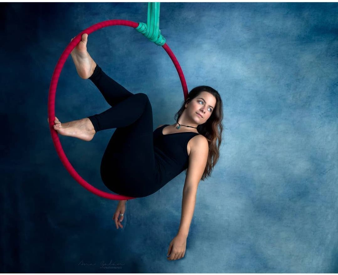 Mujer con malla negra dentro de un aro aéreo rojo, sujetado por una tela verde azulada. Fondo azul.