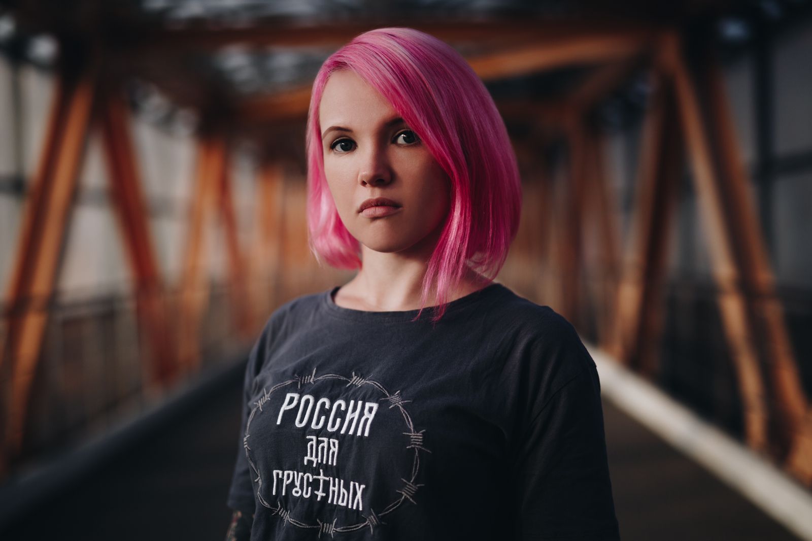 Mujer con cabello rosa, con una camisa gris con texto cirílico, parada frente a un puente de metal.