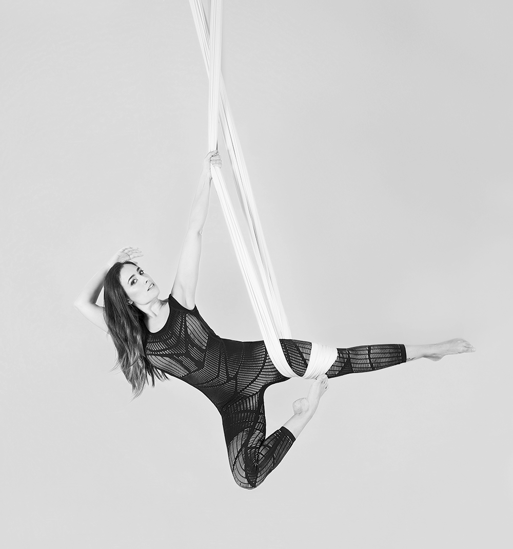 Mujer con traje aéreo negro actuando sobre tela de seda blanca, con la pierna extendida.