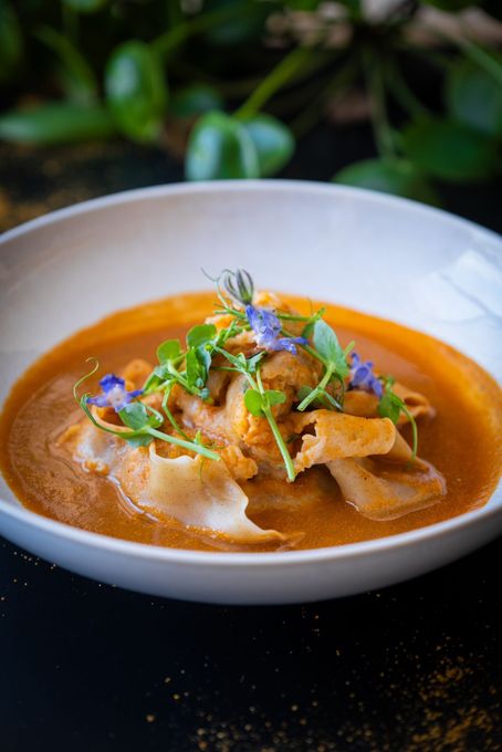 Bol blanc de soupe au curry rouge garni d'herbes et de fleurs violettes sur une table sombre.