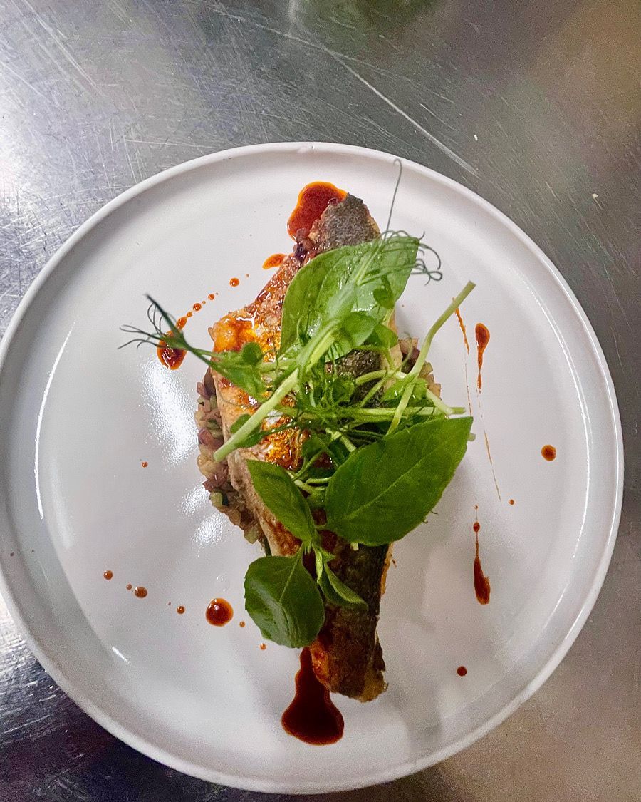 Plat gastronomique composé de légumes verts et de sauce rouge, présenté sur une assiette blanche posée sur une table en métal.