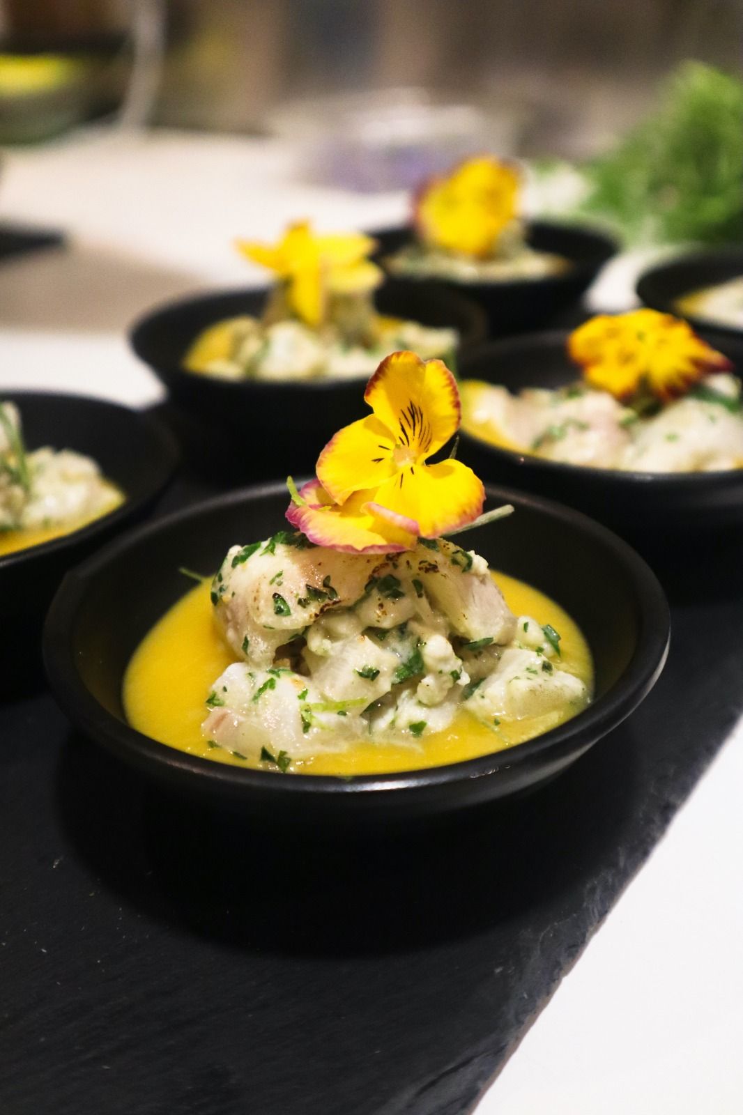 Des bols noirs contenant des mets raffinés, surmontés de fleurs comestibles jaunes, sont disposés sur une table sombre.