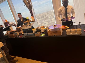 Un buffet garni de pâtisseries et de boissons est dressé à côté de deux serveurs dans une salle de conférence lumineuse donnant sur la ville.