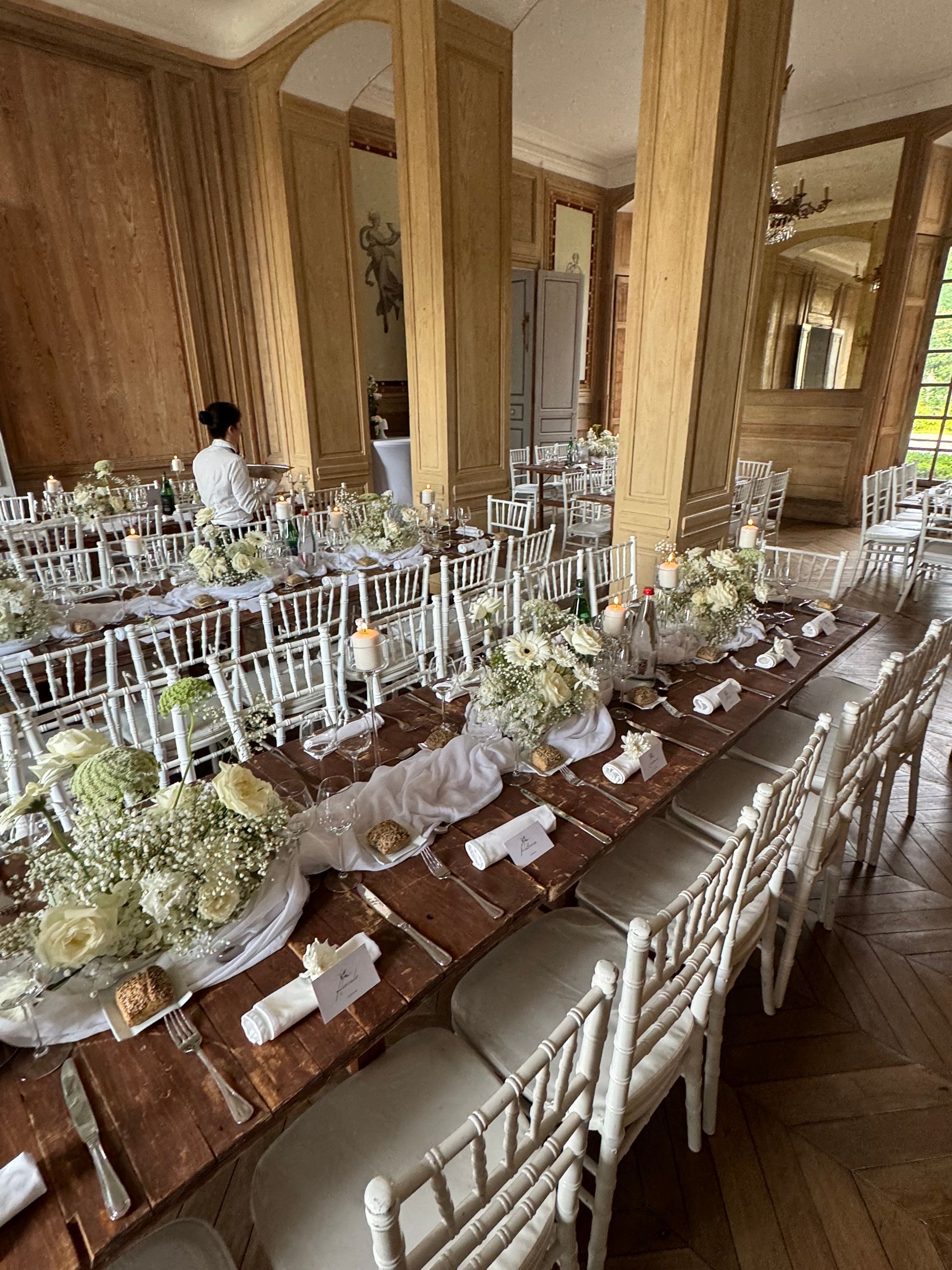 Longue table de banquet dressée avec des fleurs blanches et des chaises argentées dans une salle à manger lambrissée.
