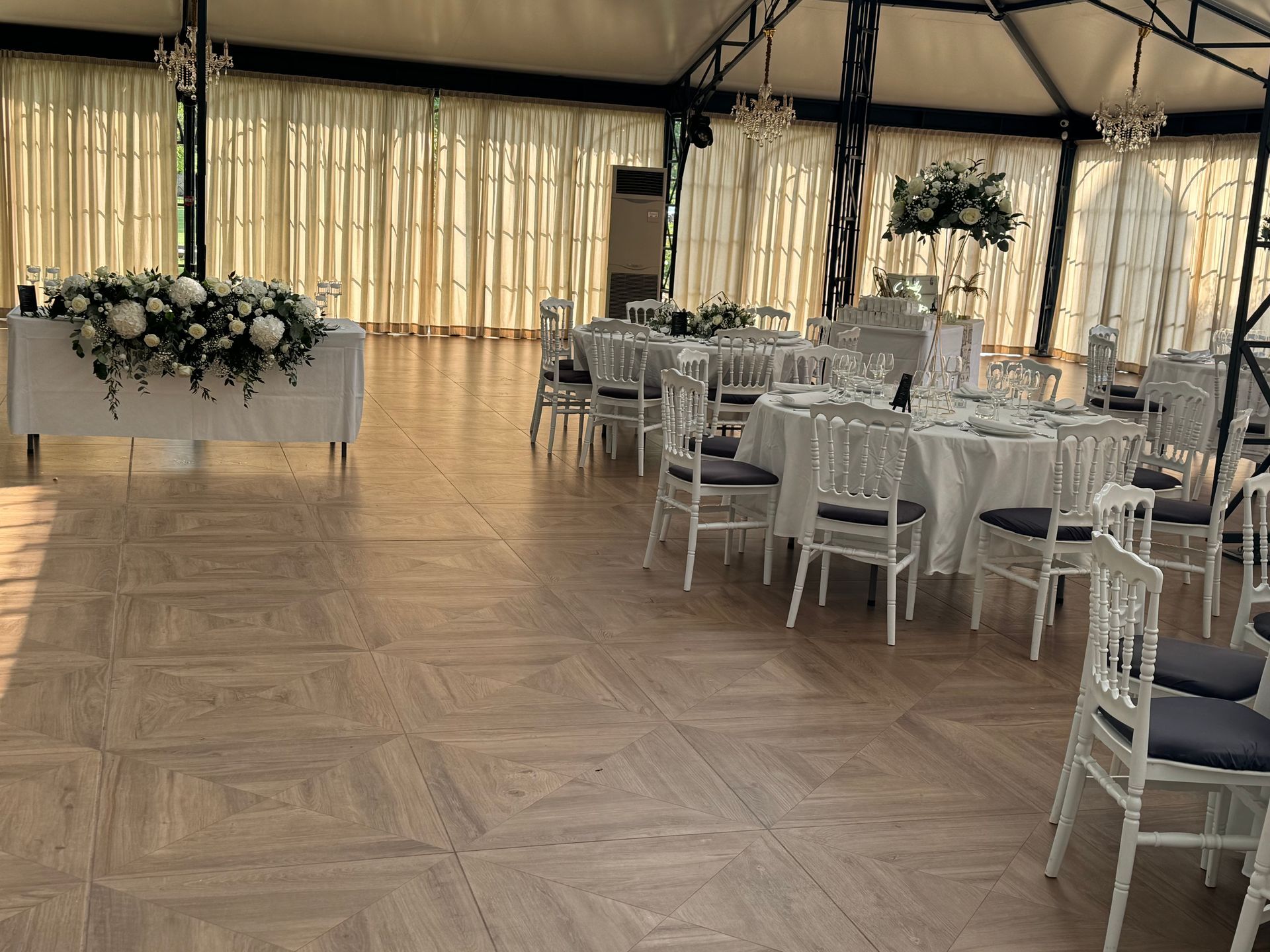 Élégante salle de banquet avec chaises blanches, tables rondes et centres de table floraux sous des rideaux drapés.