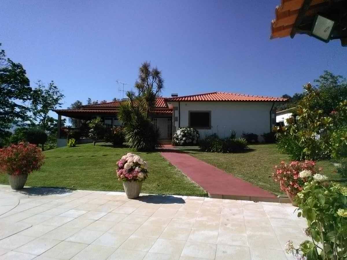 Casa con techo rojo, paredes blancas y pasarela roja; rodeada de césped verde y flores en macetas en un día soleado.