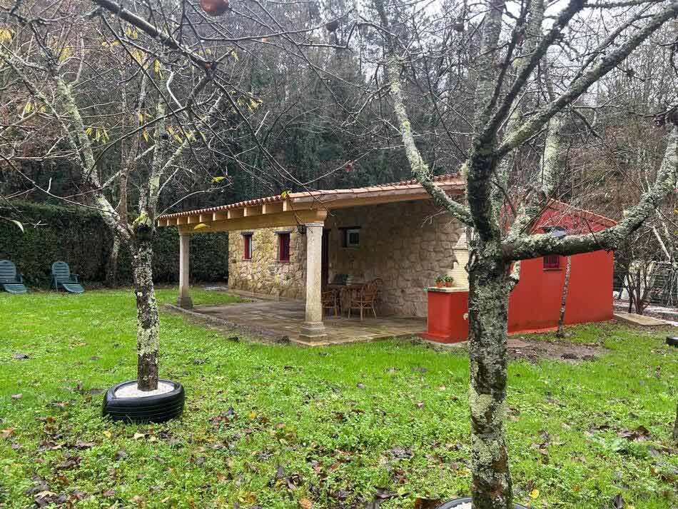 Cabaña de piedra con detalles en rojo, sobre un césped. Árboles desnudos enmarcan la escena; sillas verdes cerca.