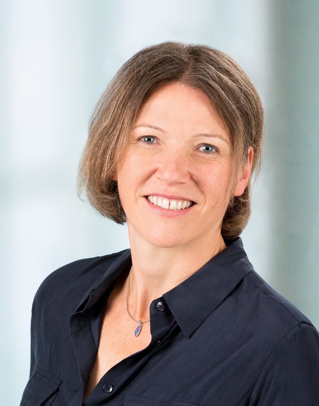 Frau mit braunen Haaren und blauer Bluse lächelt in die Kamera. Der Hintergrund ist in Blau- und Weißtönen verschwommen.
