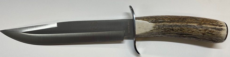 Un cuchillo de caza con hoja dentada y mango de asta yace sobre una superficie blanca.