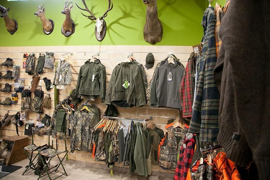 Interior de tienda de artículos de caza con chaquetas, ropa de camuflaje, accesorios de caza y cabezas de ciervo montadas.