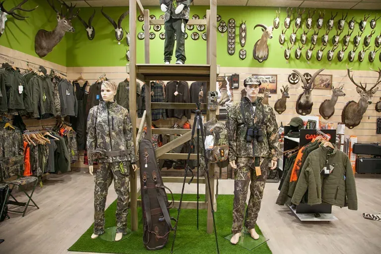Interior de tienda de ropa de caza con maniquíes con ropa de camuflaje y cabezas de animales montadas.