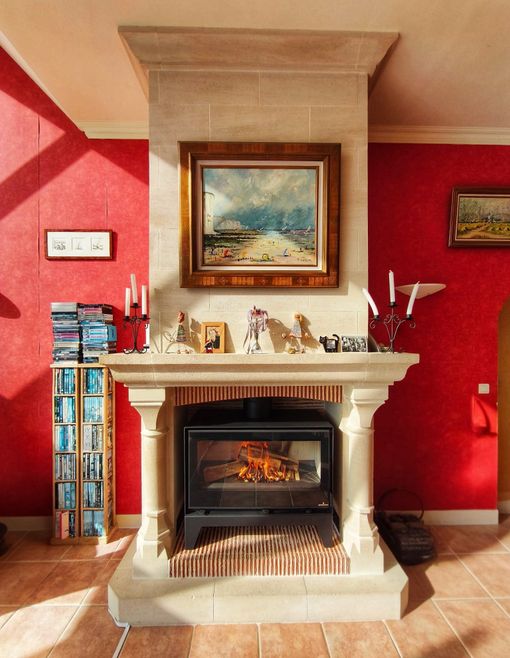 Salon rouge avec cheminée en pierre beige, tableaux encadrés et cheminée électrique allumée.