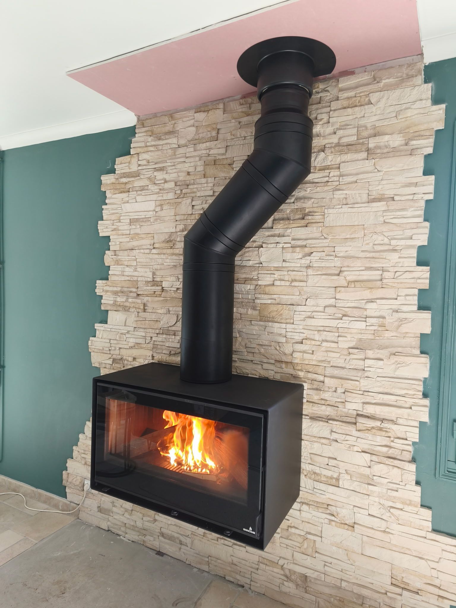 Cheminée noire moderne avec flamme visible et conduit de ventilation noir, adossée à un mur en pierre.