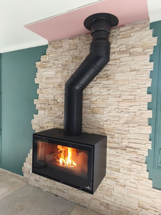 Cheminée noire moderne avec flamme visible et conduit de ventilation noir, adossée à un mur en pierre.