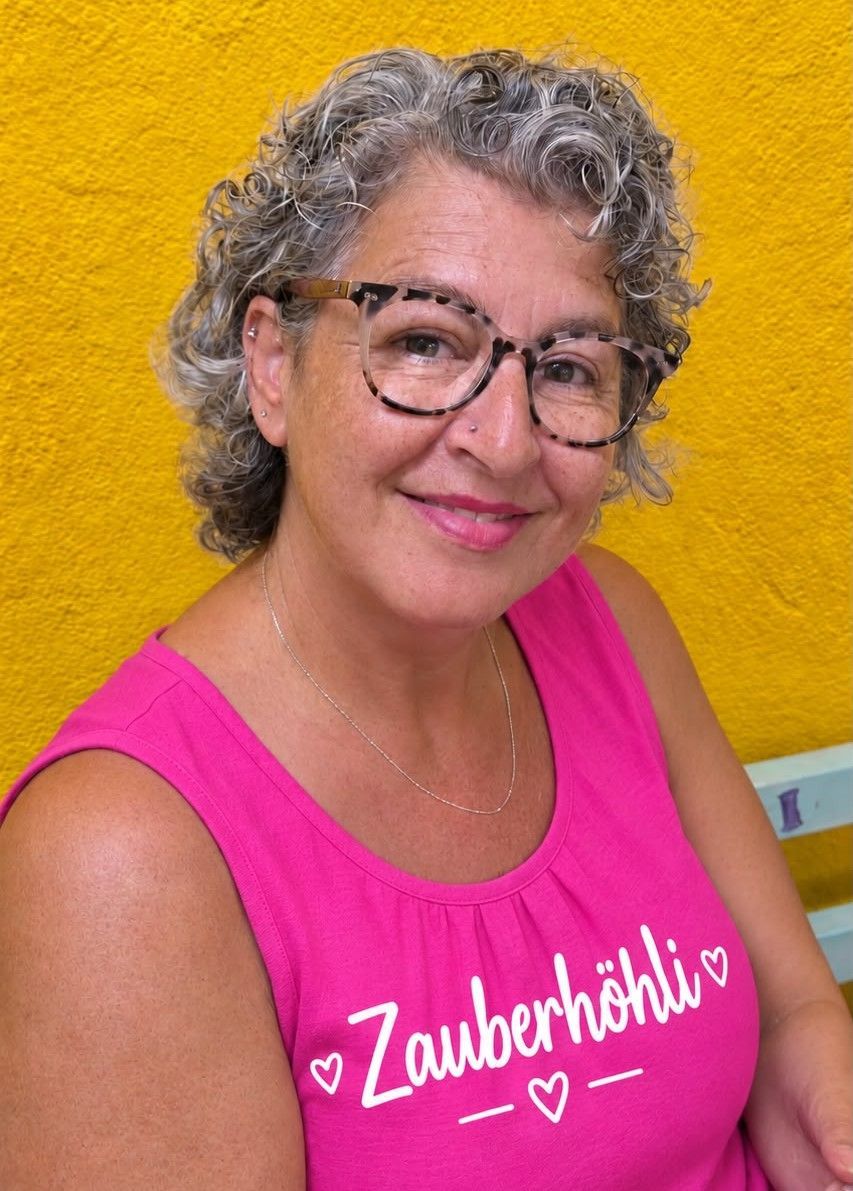 Eine lächelnde Frau mit Brille steht vor einem farbenfrohen Landschaftsgemälde.