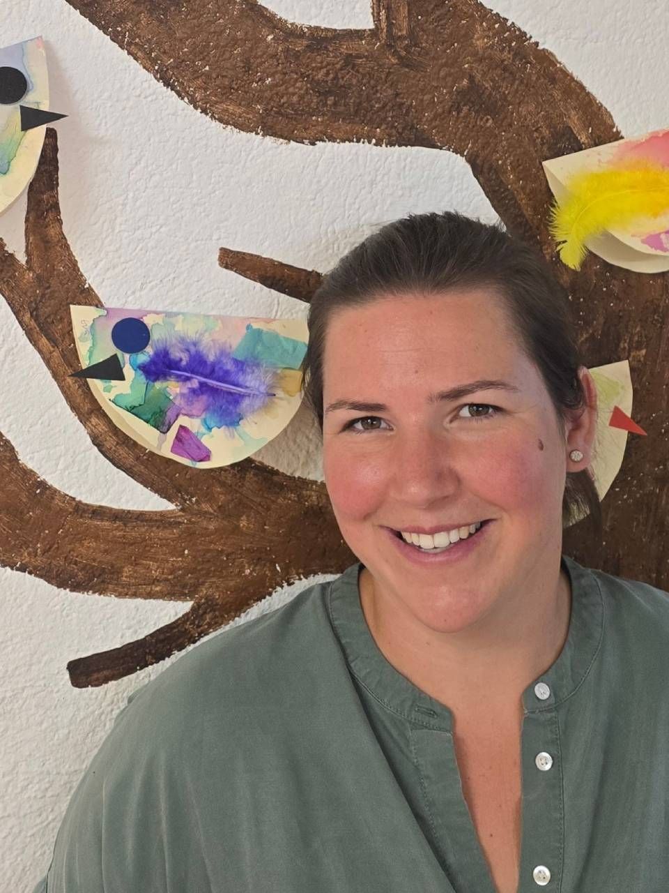Lächelnde Person in einem grünen Hemd vor einem gemalten Baum-Wandbild mit bunten Schmetterlingsverzierungen.