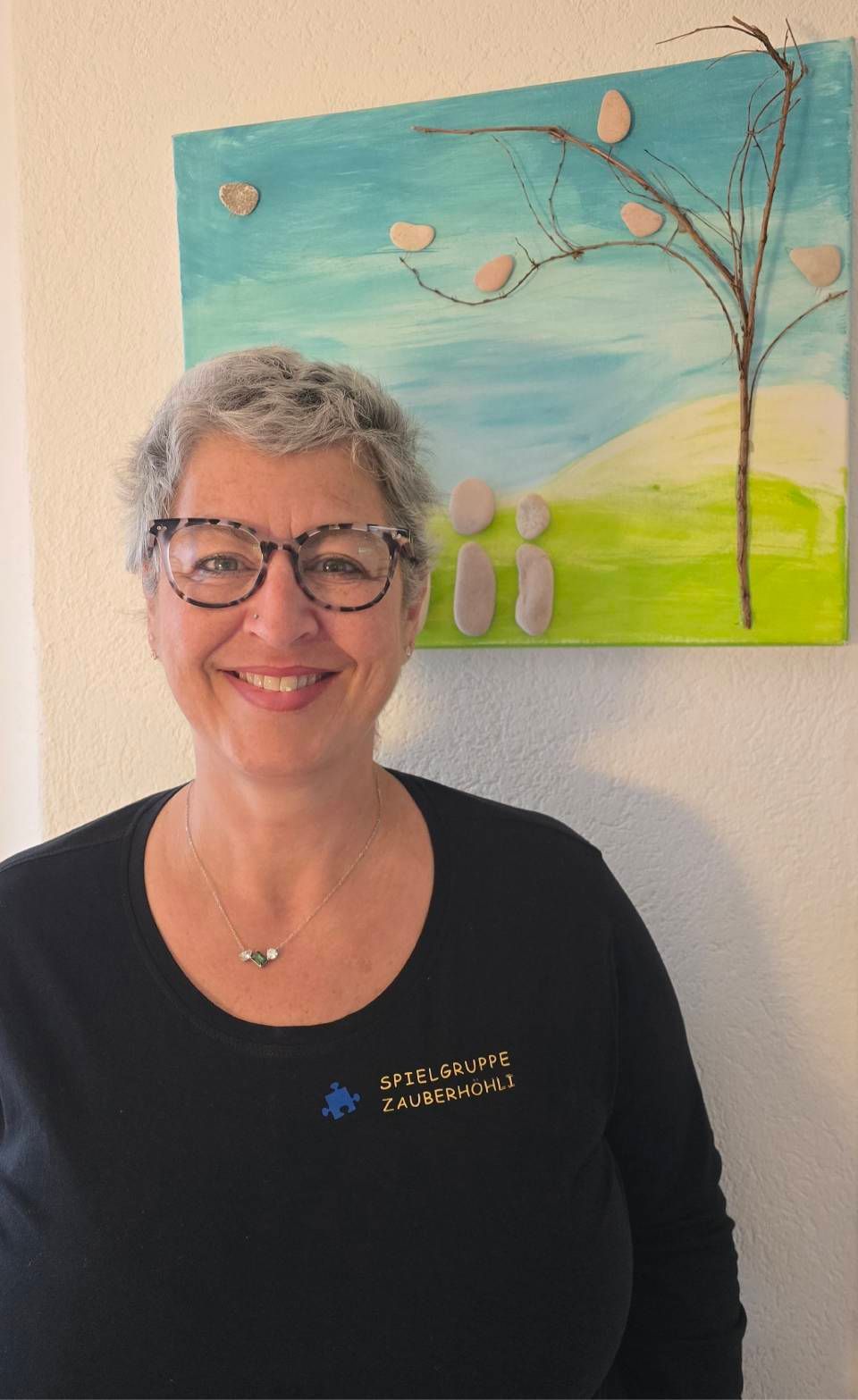 Eine lächelnde Frau mit Brille steht vor einem farbenfrohen Landschaftsgemälde.