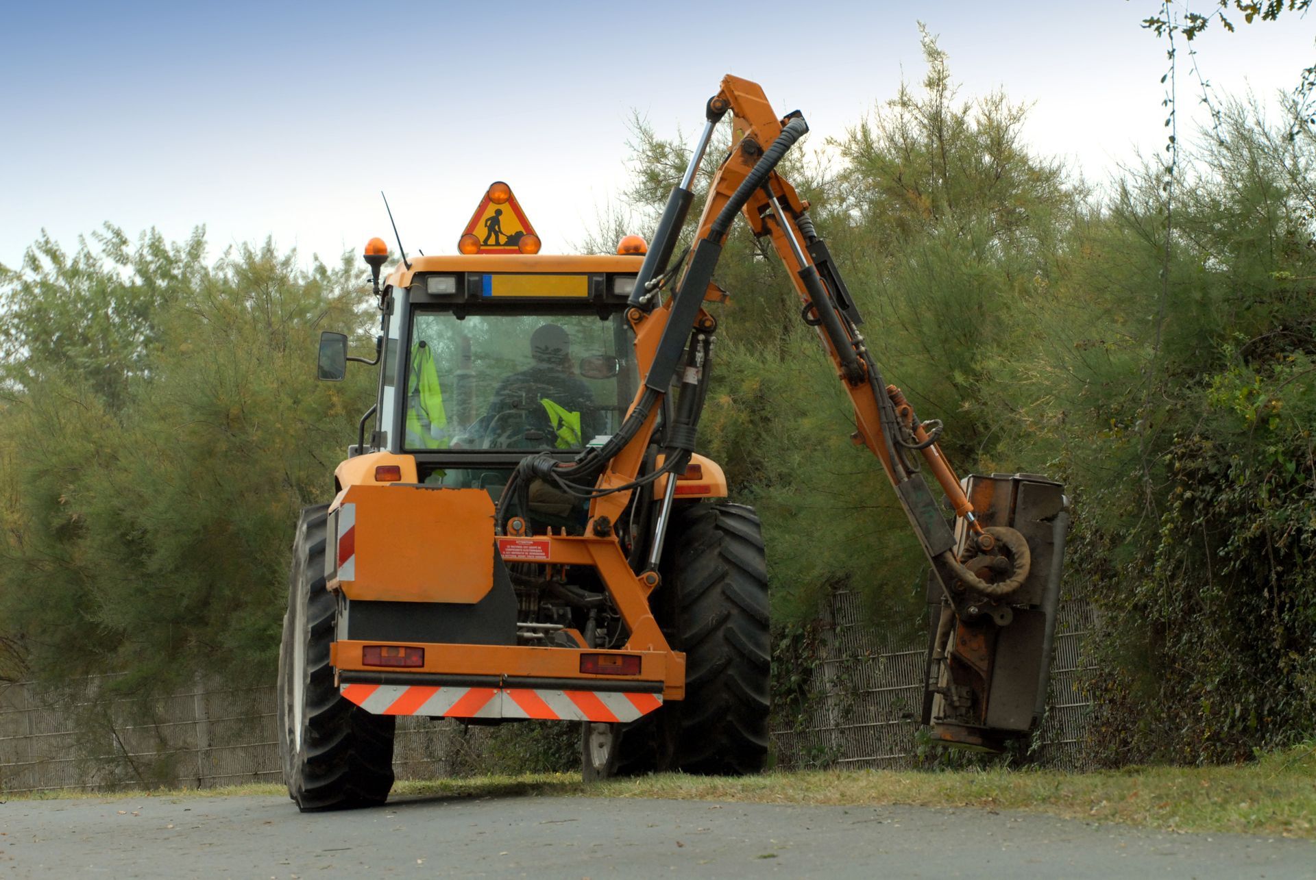 Tracteur orange avec tondeuse intégrée, taillant les buissons en bord de route.