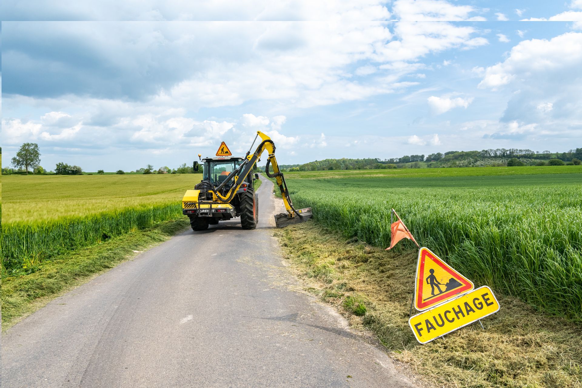 Tonte de l'herbe en bord de route : un tracteur jaune tond la pelouse au bord d'une route, un panneau d'avertissement indiquant fauchage (tonte).