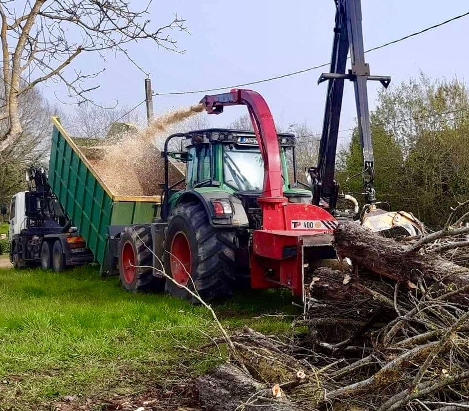 Tracteur équipé d'une déchiqueteuse à bois traitant des grumes et chargeant les copeaux de bois dans une remorque verte.