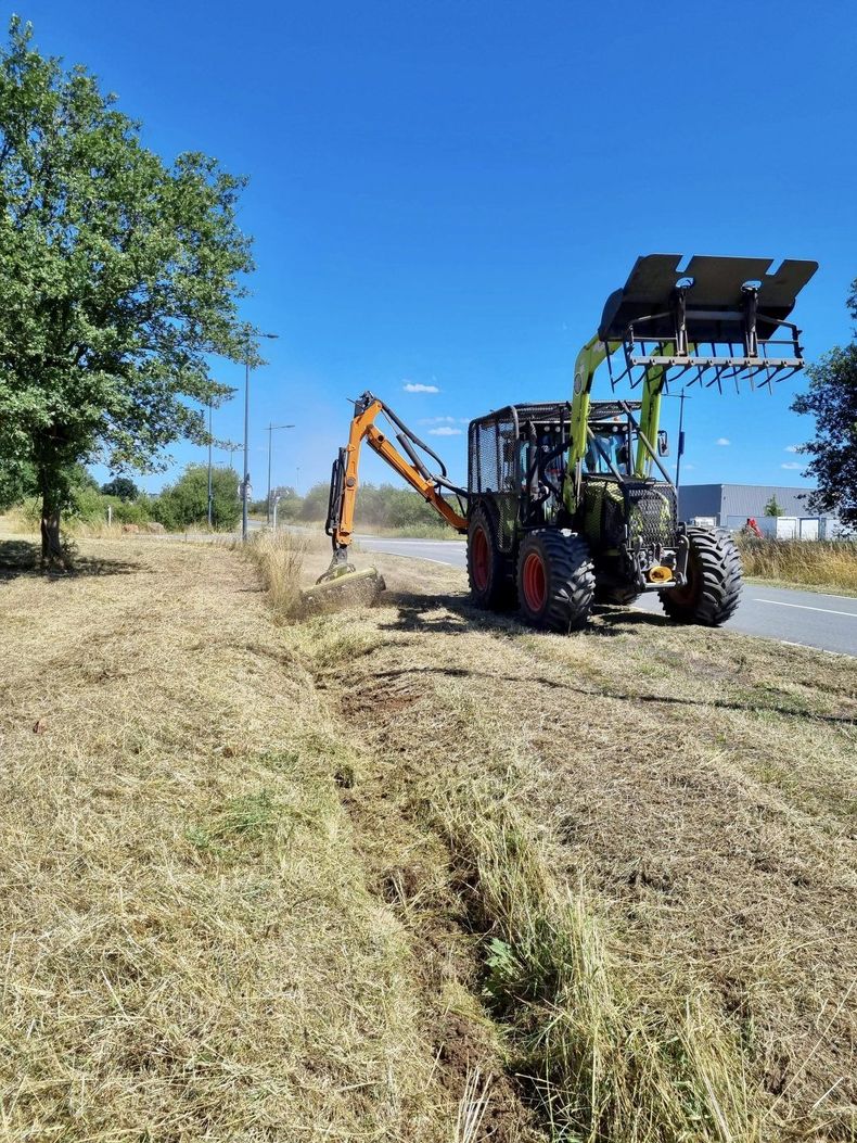 Un tracteur équipé d'un bras excavateur dégage l'herbe sèche d'un fossé en bord de route, sous un ciel bleu.