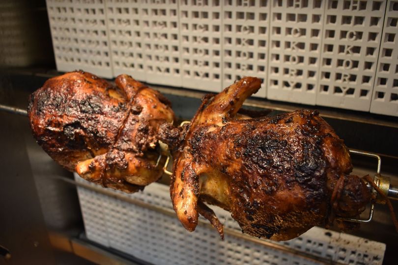 Dos pollos asados ​​girando en un asador, con un exterior marrón y crujiente, frente a un fondo de cocina comercial.