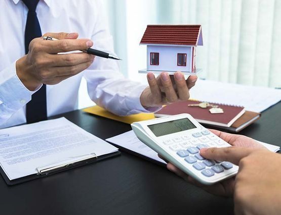 Una persona sostiene una maqueta de una casa y señala con un bolígrafo; otra usa una calculadora; hay papeles y llaves sobre el escritorio.