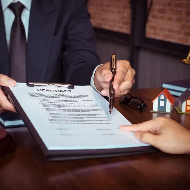 Persona firmando contrato, bolígrafo en mano, mientras otra persona señala. Se ven maquetas de casas.