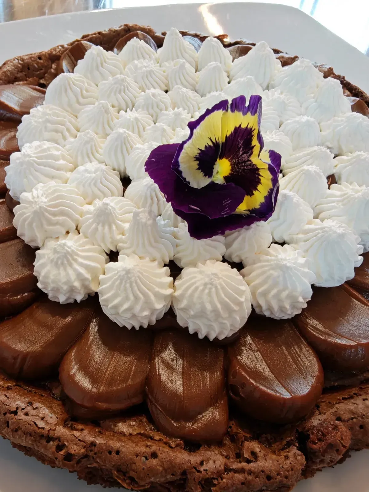 Pastel de chocolate decorado con rosetas de crema batida blanca y una flor comestible de color amarillo púrpura.