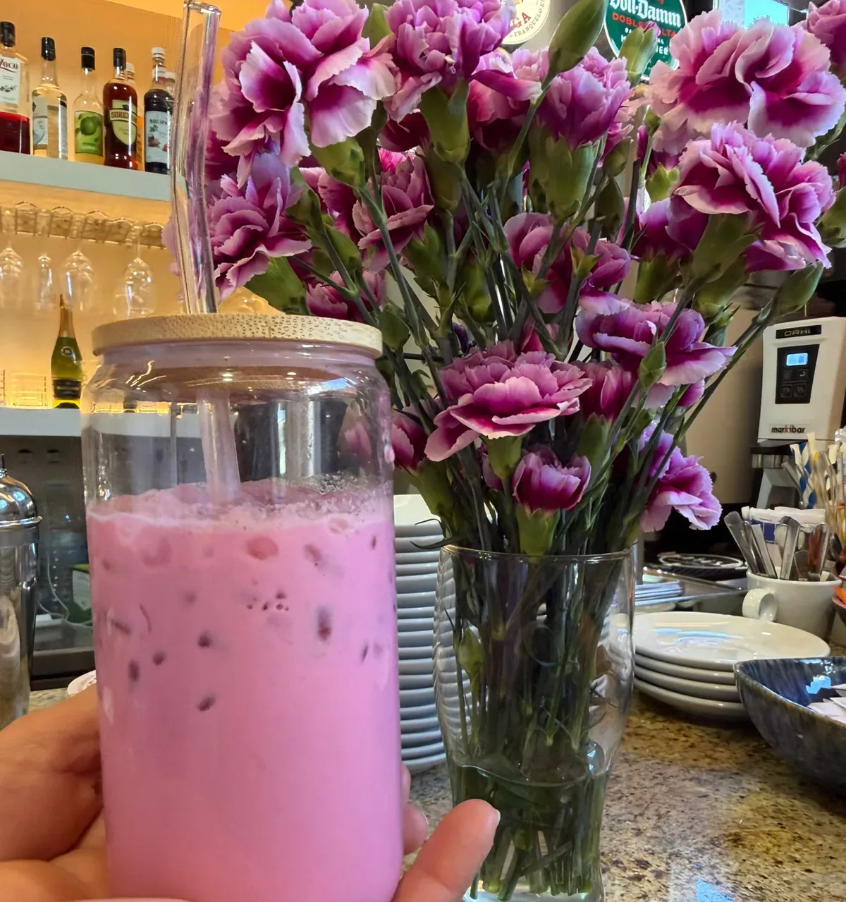 Bebida rosa helada en un vaso junto a un jarrón de flores moradas en la barra de una cafetería.