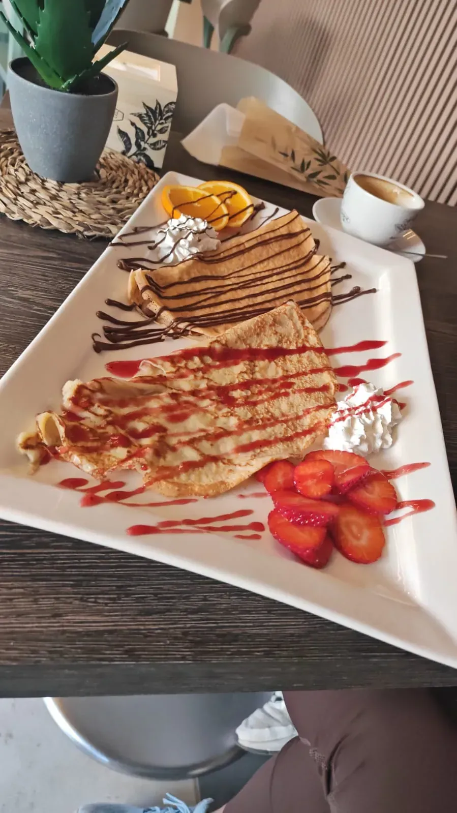 Crepe de postre en un plato cuadrado con sirope de chocolate, fresas y nata montada sobre una mesa.