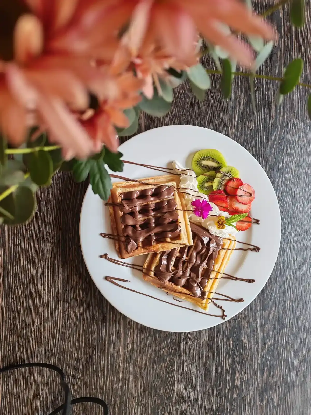 Gofres con sirope de chocolate y fruta en un plato blanco, con flores difuminadas en primer plano.