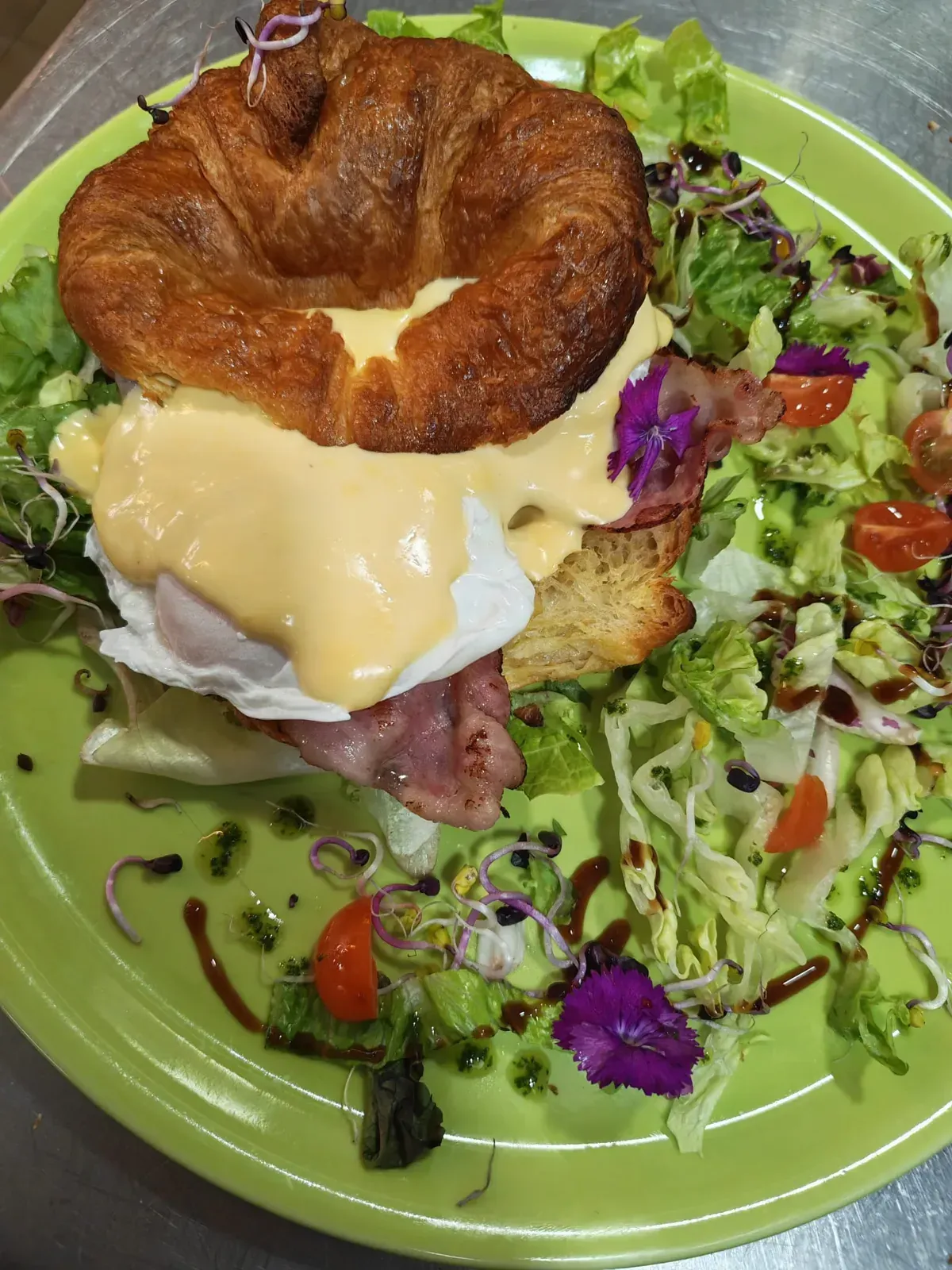 Sándwich abierto con huevo frito y queso fundido en un plato verde, adornado con ensalada y tomates cherry.