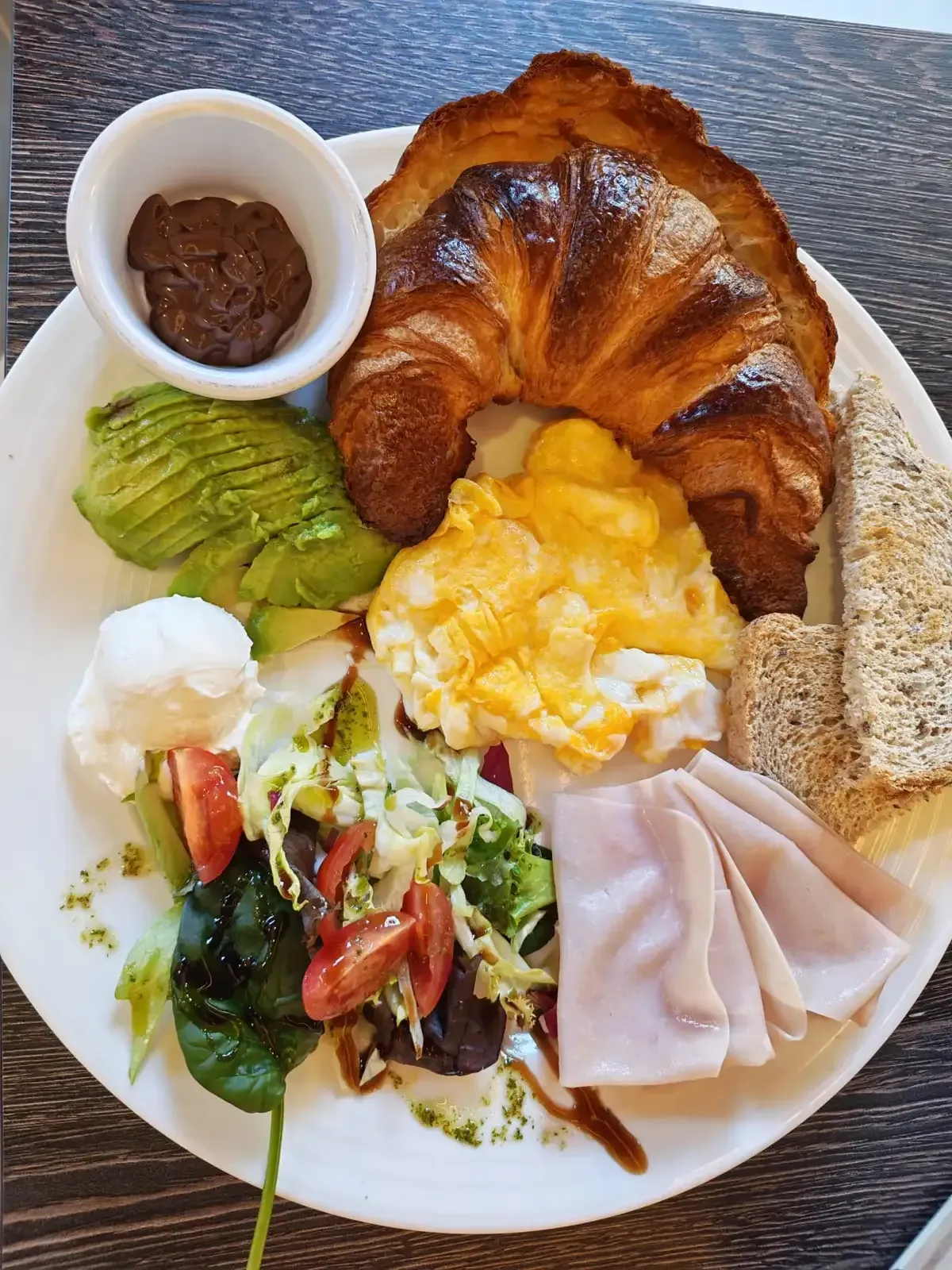 Plato de desayuno con croissant, huevos revueltos, aguacate, ensalada, jamón, pan y una pequeña porción de untable.