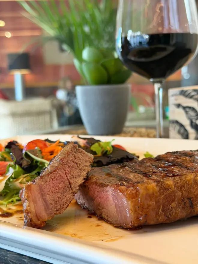 Filete servido con verduras en la mesa de un restaurante, con una copa de vino tinto y una planta borrosa al fondo.