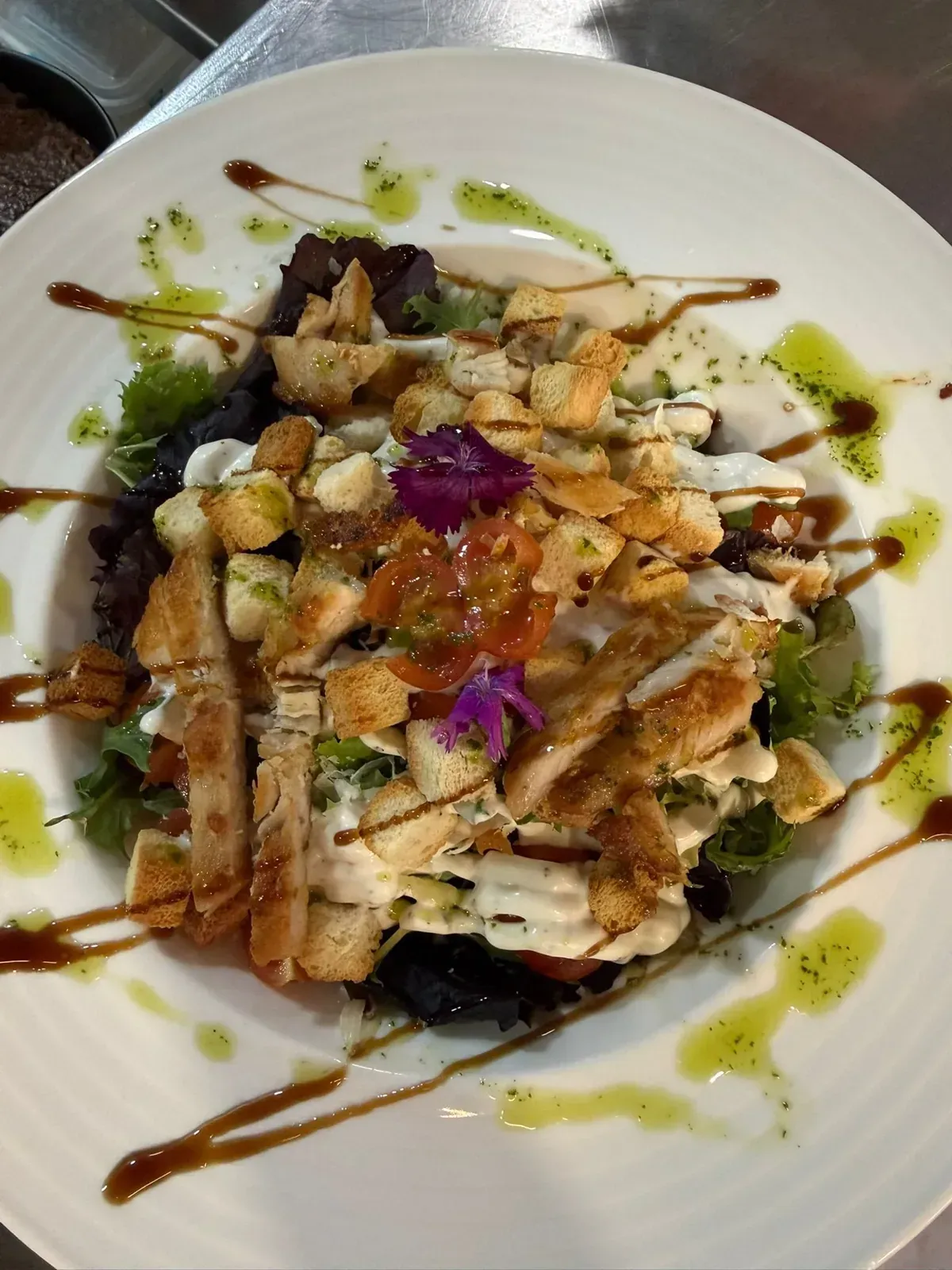 Ensalada de pollo a la parrilla emplatada con verduras, salsa y flores comestibles moradas en un plato blanco.