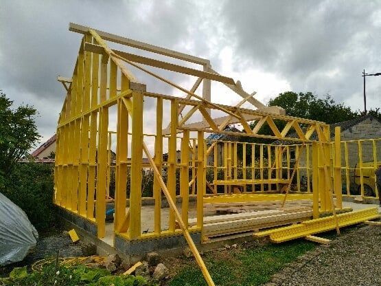Ossature bois d'une maison en construction, vue extérieure. Bois jaune, ciel nuageux, herbe et fondations en pierre.