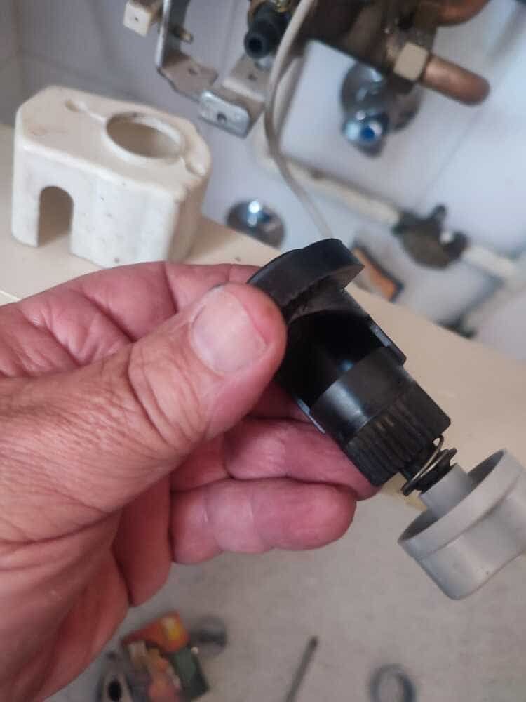 Mano sosteniendo un accesorio de plomería de plástico negro con una tapa gris cerca de un conjunto de tuberías montado en la pared.