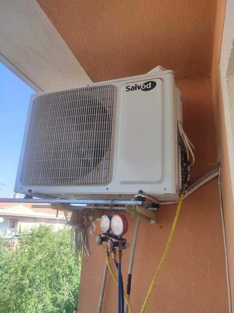 Unidad exterior de aire acondicionado montada en una pared bajo un alero, con mangueras y tuberías conectadas.