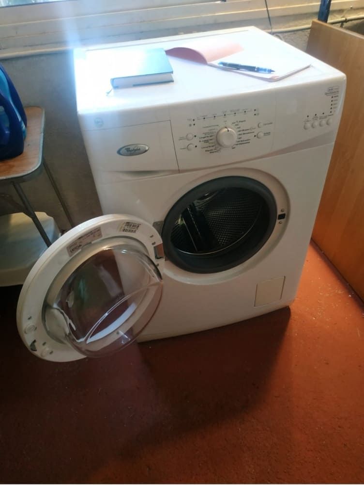 Lavadora de carga frontal con la puerta abierta, electrodoméstico blanco para lavandería sobre suelo naranja junto a una ventana.