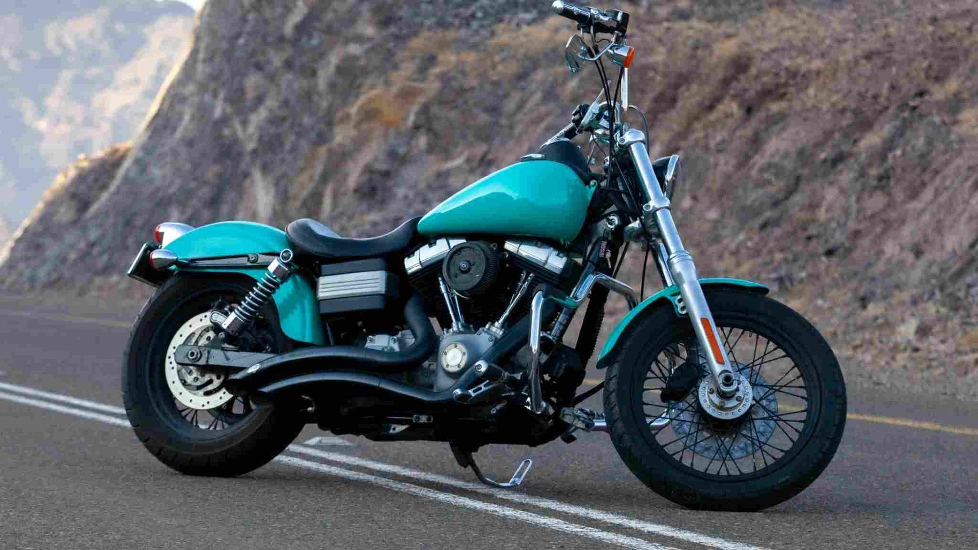 Motocicleta Harley-Davidson color verde azulado en una carretera pavimentada con un fondo de montañas.