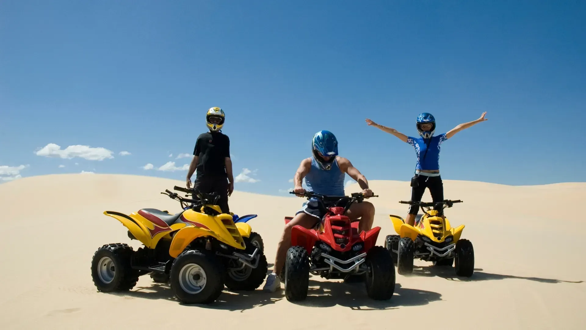 Tres personas en vehículos todoterreno en un entorno desértico con cielo azul. Una de pie, dos conduciendo, todas con casco.