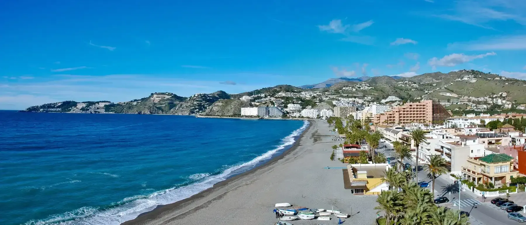 Pueblo costero con una playa curva, océano azul, edificios en la ladera y coches aparcados a lo largo de la costa.