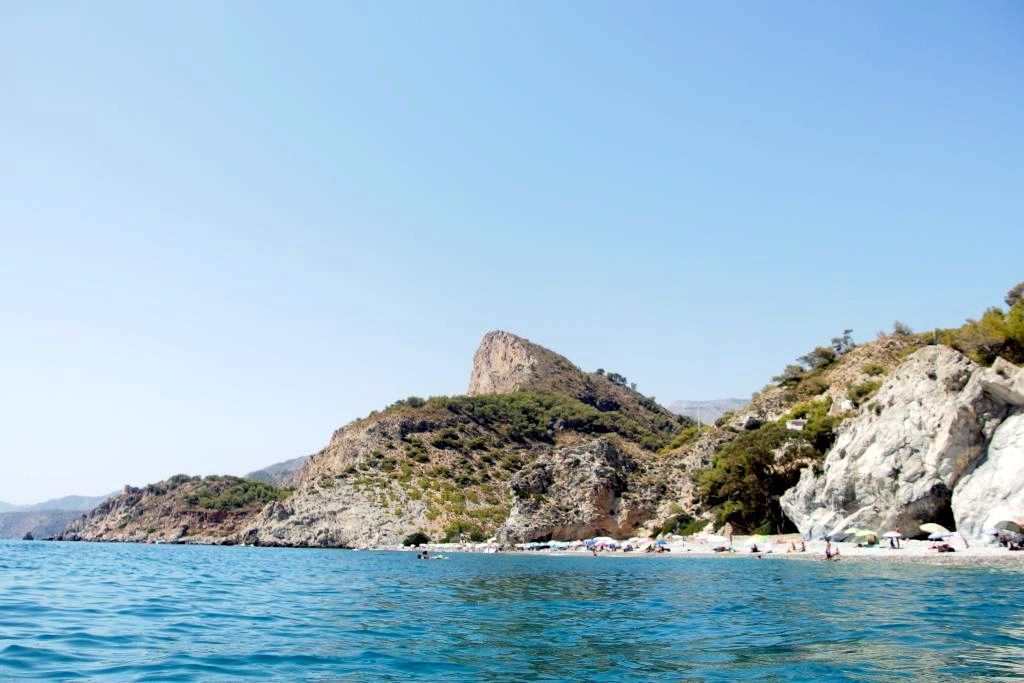 Cala rocosa costera con aguas turquesas, una pequeña playa y una colina bajo un cielo azul claro.