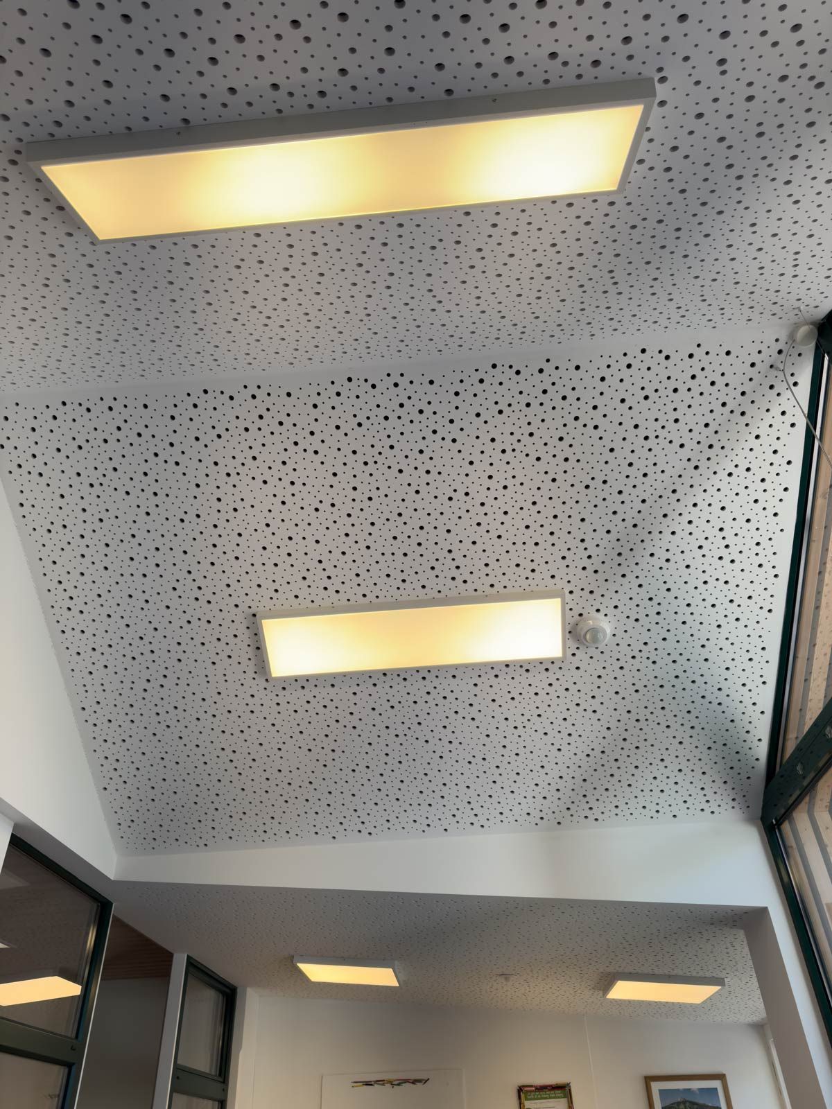 Perforierte weiße Decke mit eingelassenen rechteckigen Leuchten über einem Flur und einer Glaswand